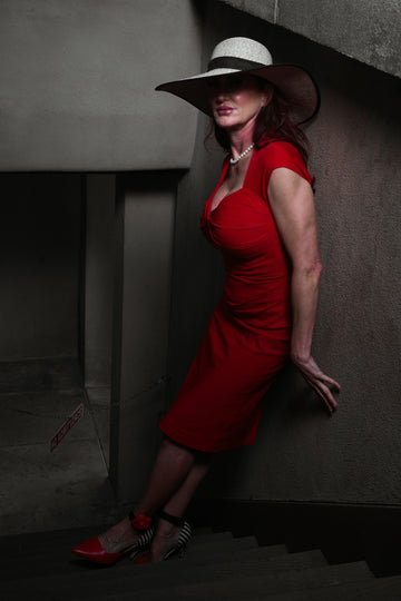 A woman in a red dress and hat leaning against a wall