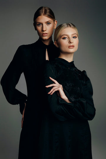 Two women in black dresses posing against a grey background.