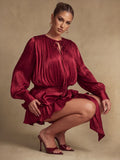 Seated view of a burgundy pleated mini dress showing its tie neckline, gathered waist, and layered ruffle skirt