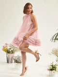 A woman models a light pink sleeveless tulle dress with a ruffled hem, paired with beige high-heeled sandals, set against a white studio background decorated with potted plants.