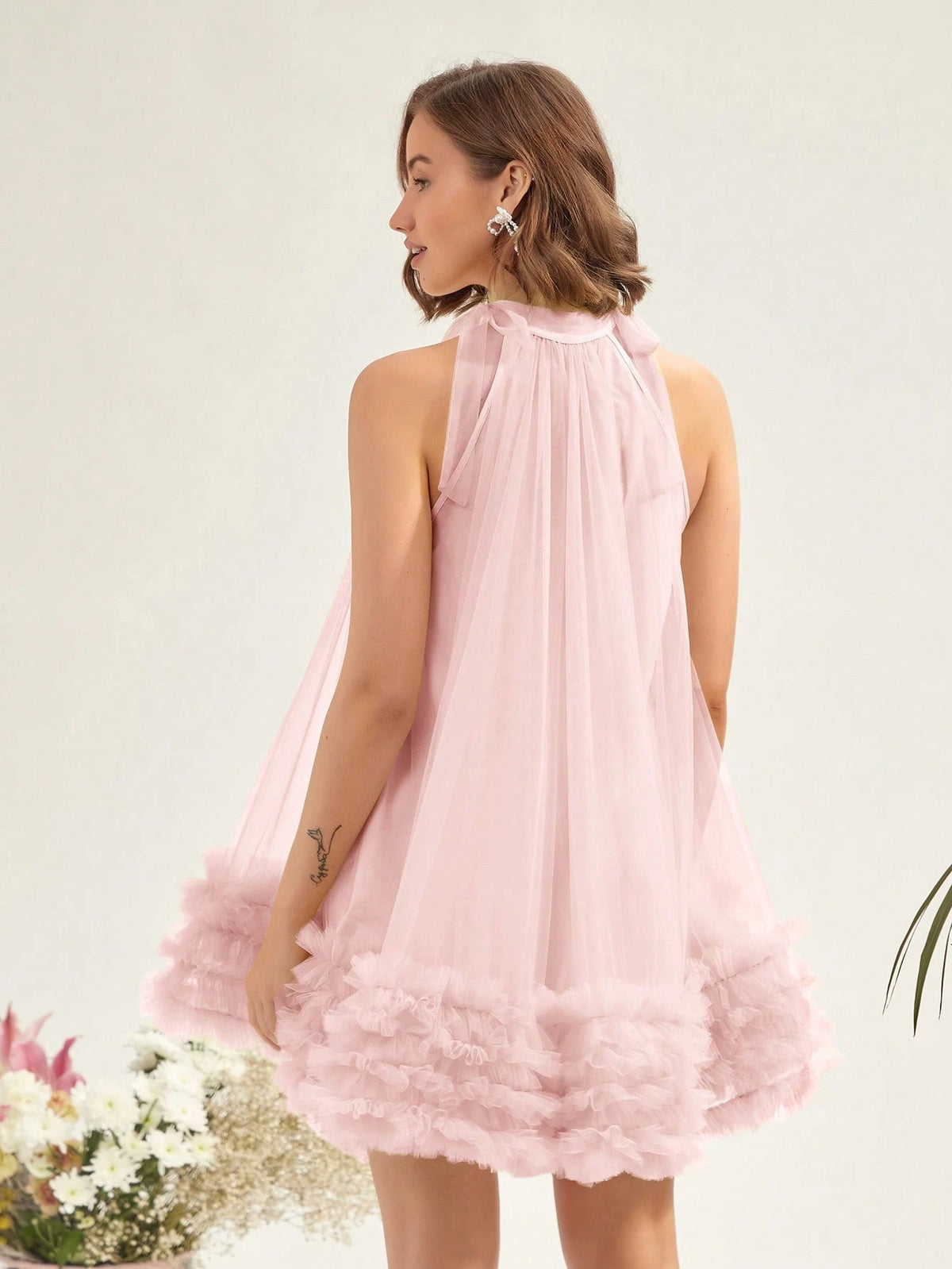 A woman wearing a light pink sleeveless tulle dress, shown from the back. The dress features a gathered halter-style neckline and a layered, ruffled tulle hem.