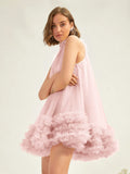 A woman wearing a sleeveless light pink dress with a high mock neck and a voluminous ruffled tulle hem, posed against a plain light background.