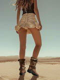 A model from behind wearing a black sleeveless top, a beige ruffled mini skirt with side buttons, and brown ankle boots, standing on sand dunes under a clear sky.