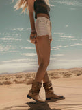 A woman standing in a desert landscape wearing light beige denim shorts, a dark crop top, and brown leather boots with buckles.