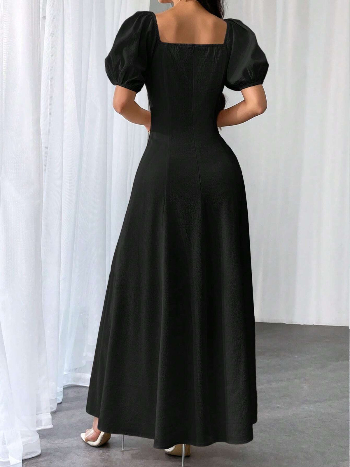 A woman wearing a long black dress with puff sleeves and a square neckline, shown from the back in a studio setting with white curtains and a gray floor.
