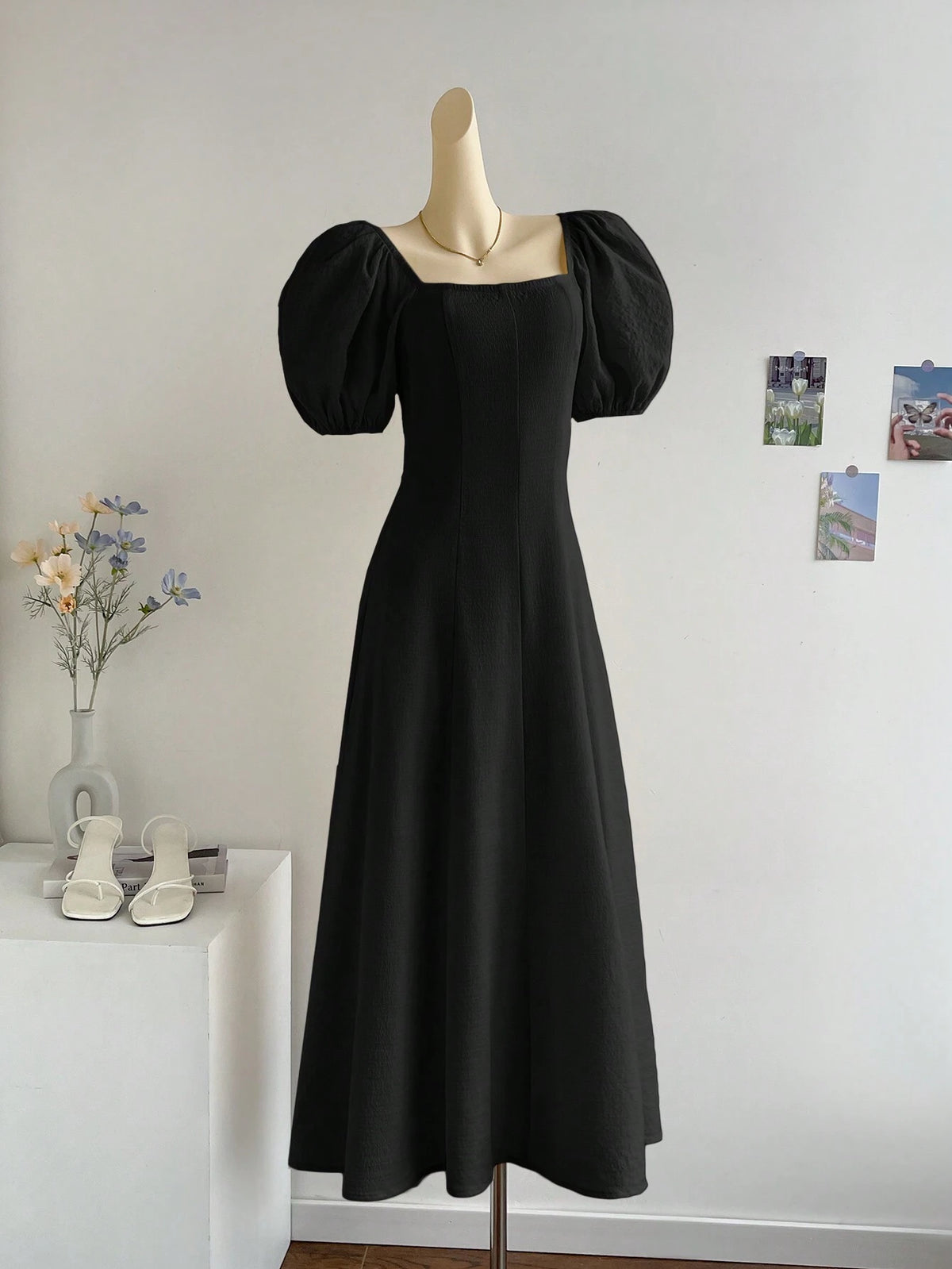 A black midi-length dress with voluminous puff sleeves and a square neckline, displayed on a mannequin in an indoor room setting with a white wall, small wall photos, a vase with flowers, and white sandals on a side table.