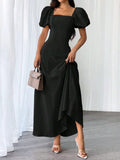 A woman models a black midi dress with puff sleeves, a square neckline, and an asymmetrical draped hem. She is holding a small taupe handbag and wearing clear-strap high heels, standing against a white curtain backdrop.