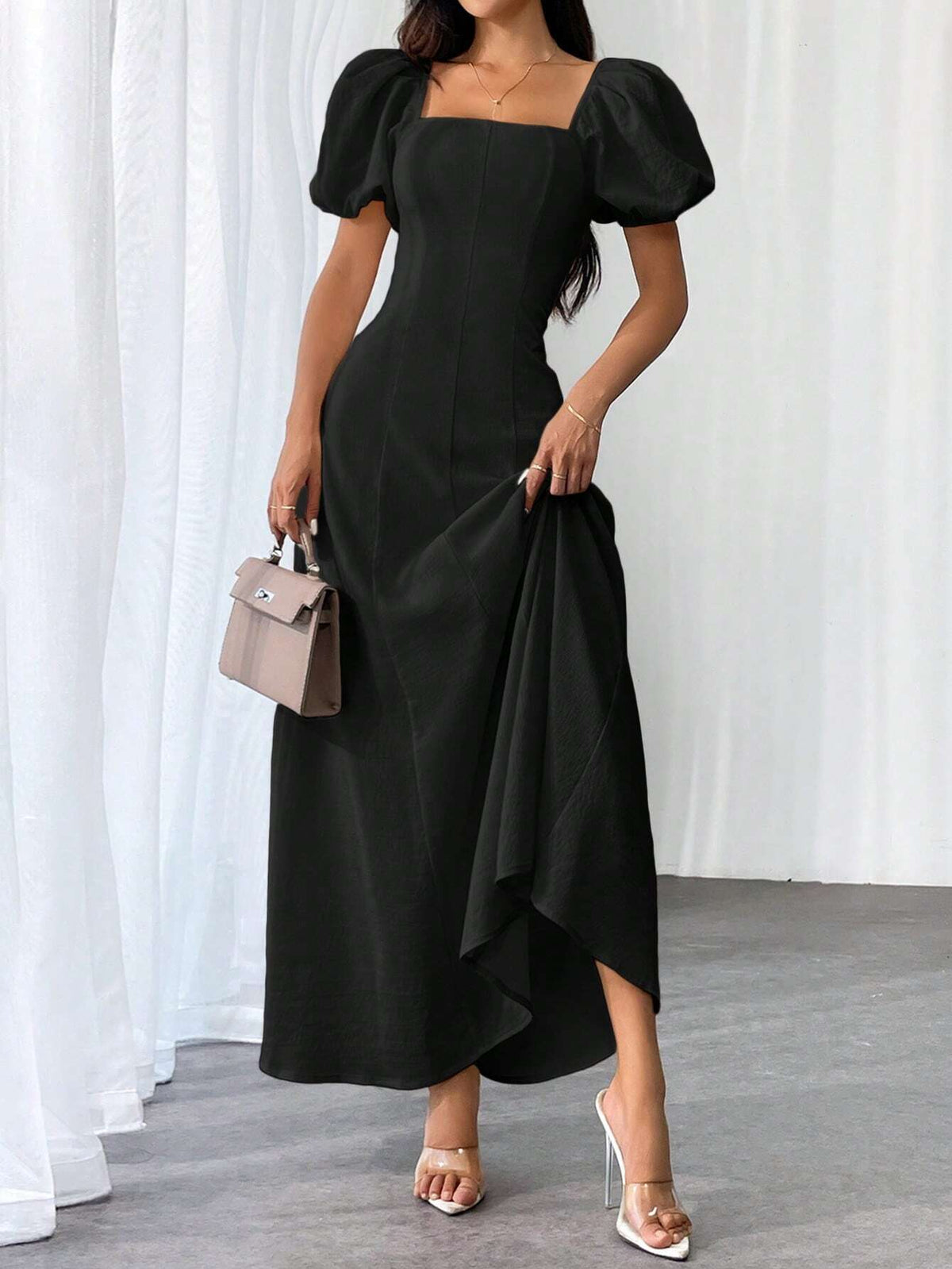 A woman models a black midi dress with puff sleeves, a square neckline, and an asymmetrical draped hem. She is holding a small taupe handbag and wearing clear-strap high heels, standing against a white curtain backdrop.