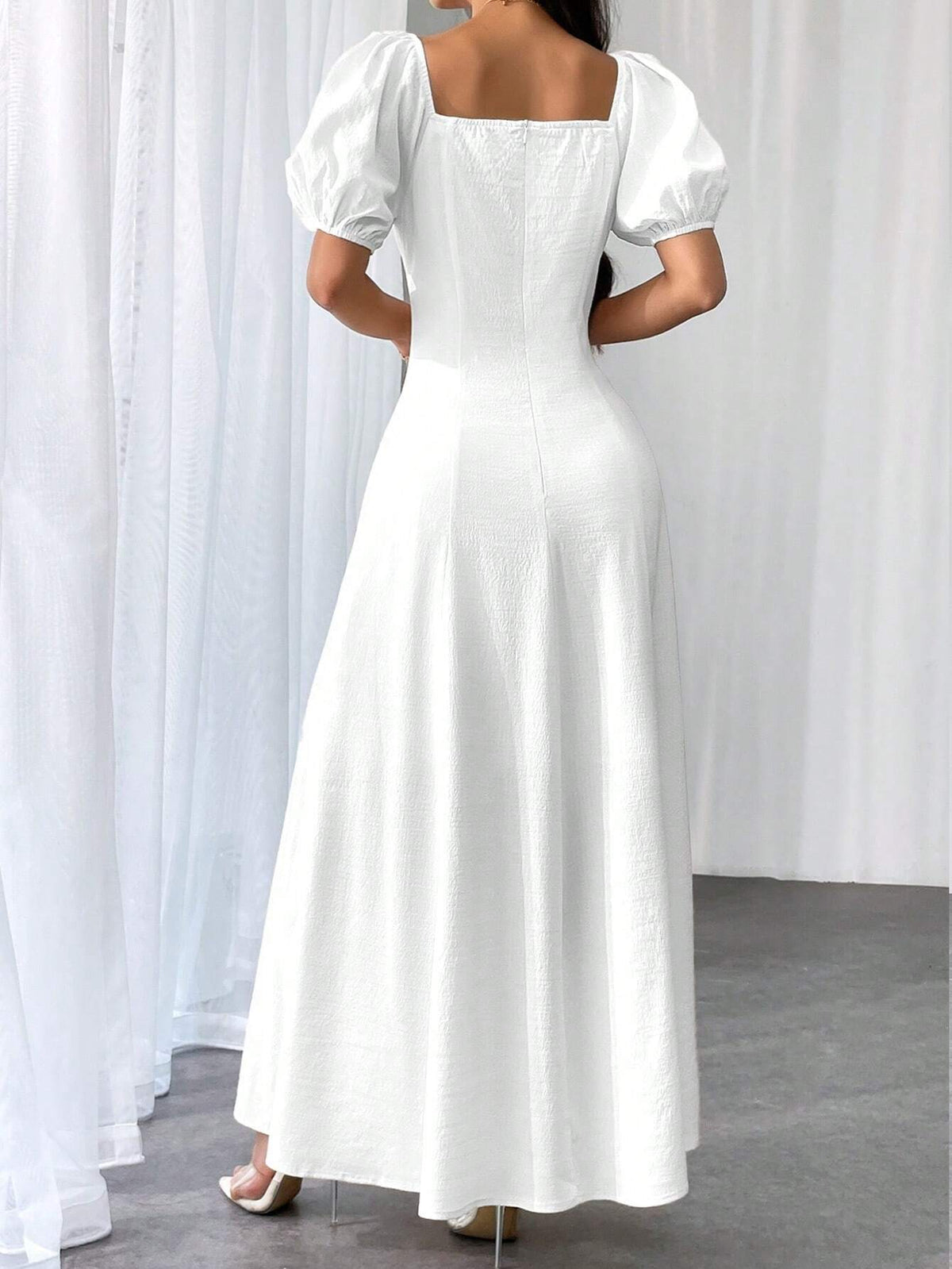 A woman wearing a white maxi dress with puff sleeves and a square neckline, shown from the back in a studio setting with white curtains and a gray floor.