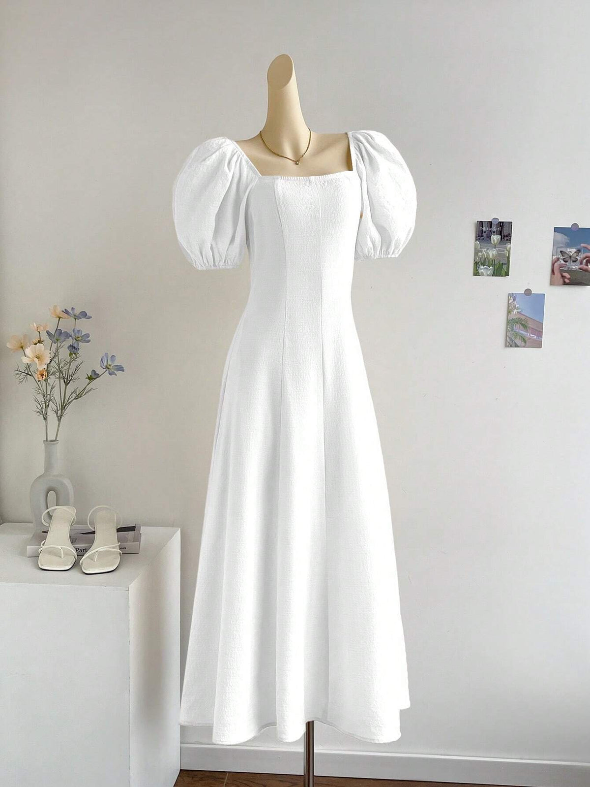 A white midi-length dress with puff sleeves and a square neckline displayed on a mannequin in a bright indoor room. The mannequin stands beside a white side table holding a vase with flowers and a pair of white sandals, with three small photos pinned to the wall behind.