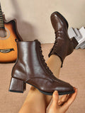 A pair of dark brown faux-leather lace-up ankle boots with block heels, shown on a model's feet and held in one hand against a neutral indoor background with a guitar and books.