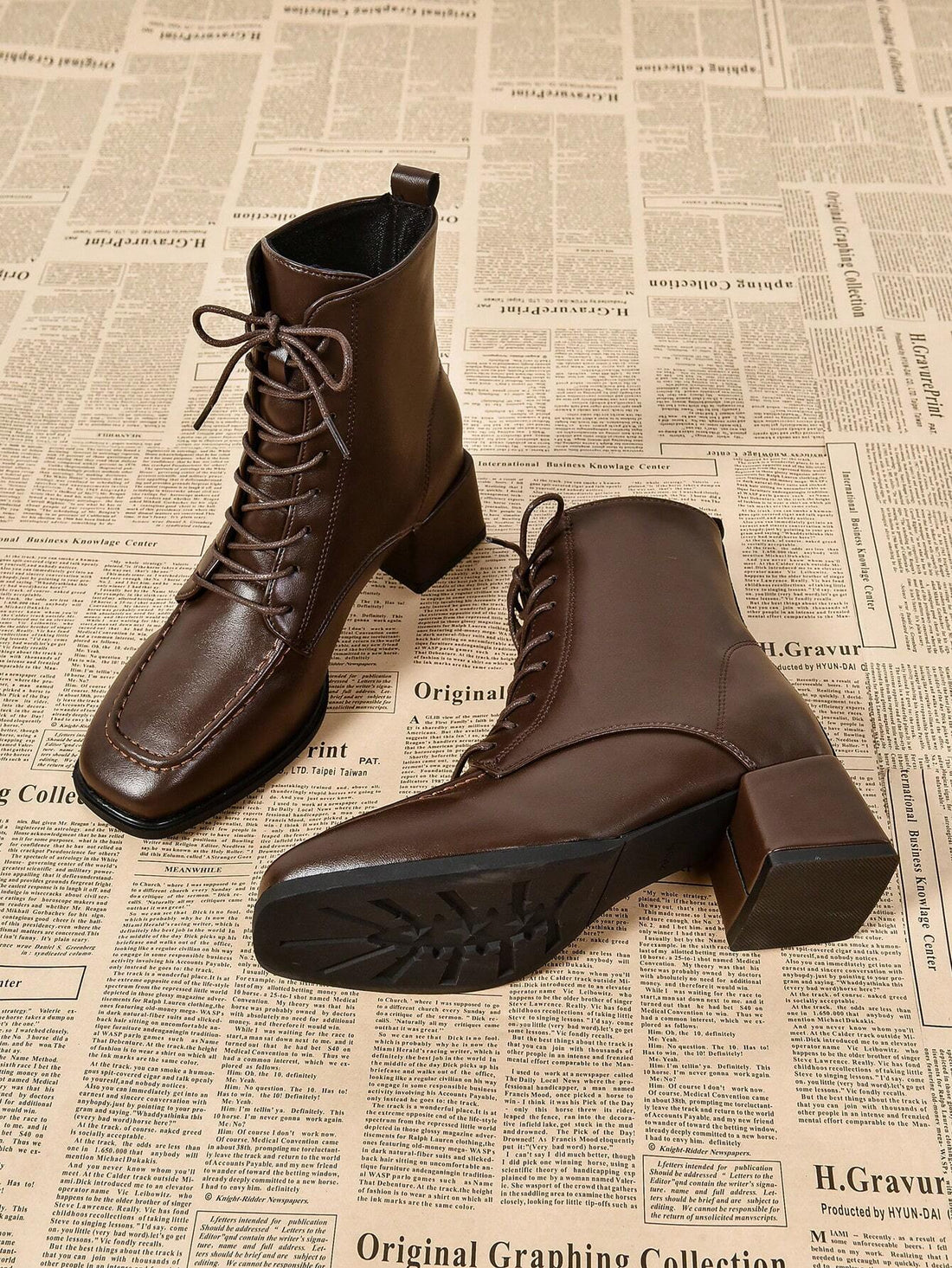A pair of dark brown leather lace-up ankle boots with block heels, shown on a model's feet against a neutral indoor background with a guitar and books.