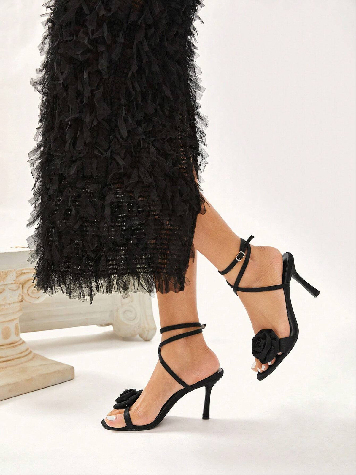 Black high-heeled sandals with floral details worn with a black ruffled dress on a white background