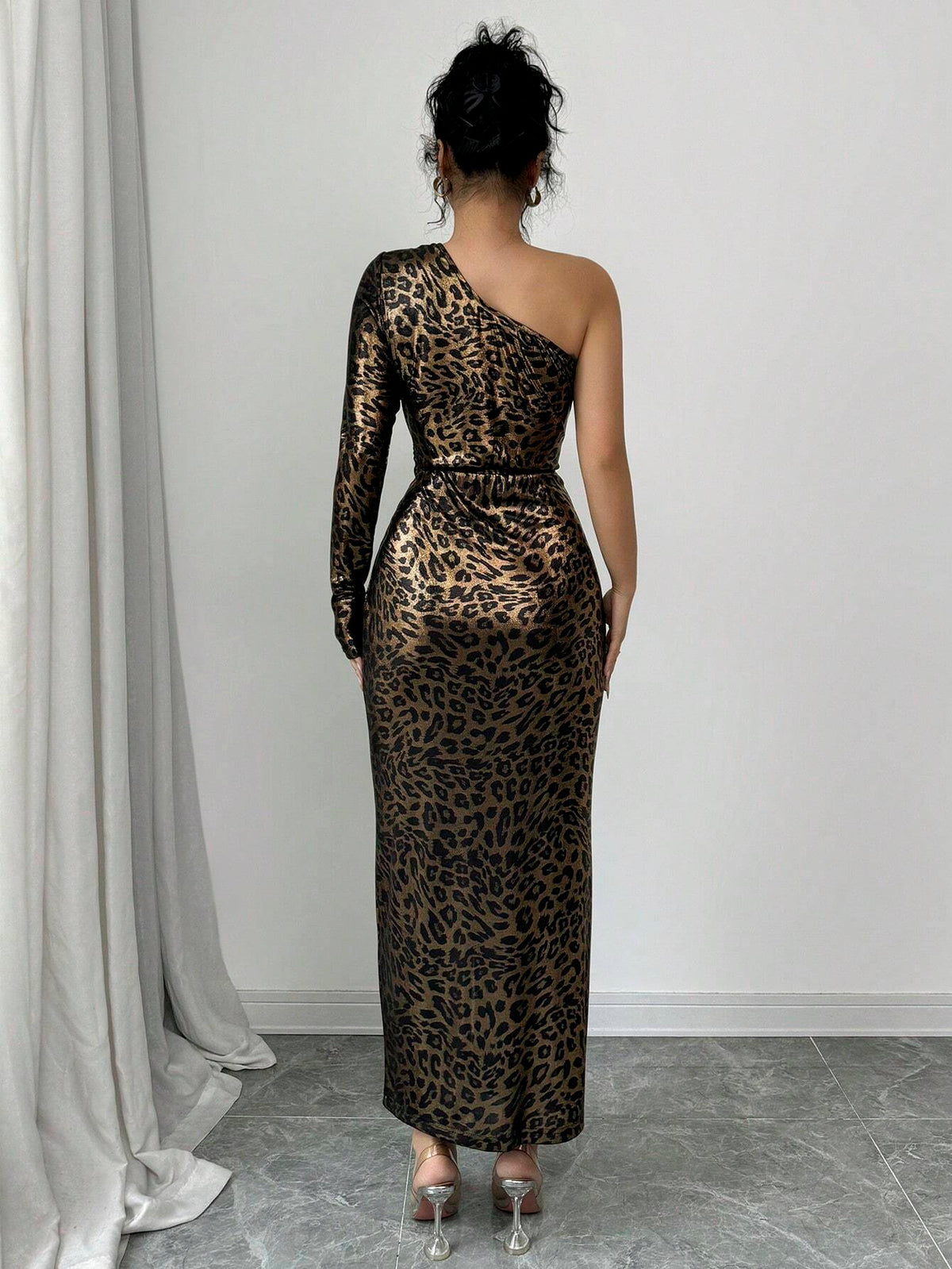 A woman viewed from behind wearing a one-shoulder leopard-print maxi dress with long sleeves and a fitted silhouette, standing in a room with a white wall, gray floor, and draped curtain.