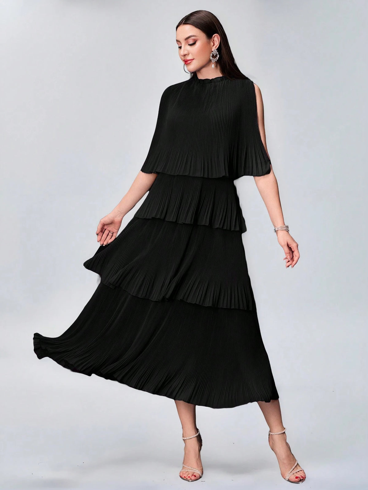 A woman modeling a black pleated midi dress with a tiered ruffle skirt, cold-shoulder sleeves, and a high neckline, posed against a light gray studio background.
