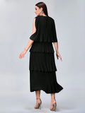 A woman modeling a black tiered pleated dress from the back, featuring a cold-shoulder design and matching pleated skirt, paired with clear high-heeled sandals against a light gray studio background.
