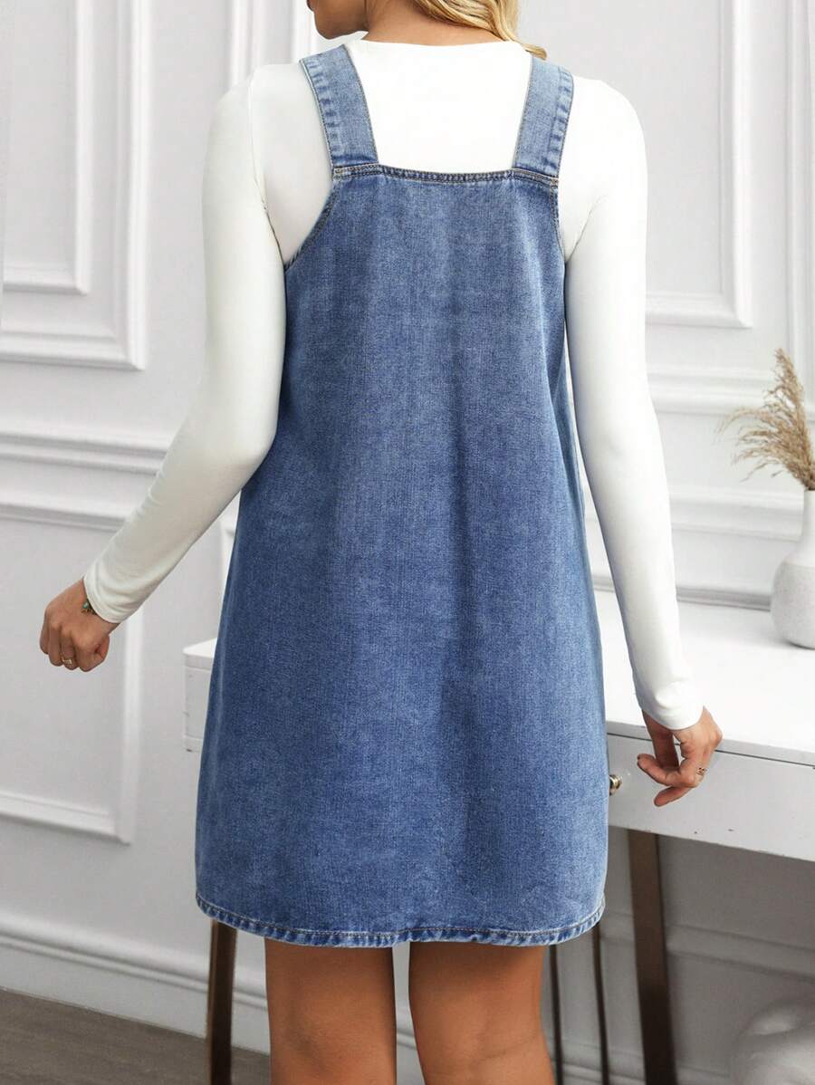 Front detail view of medium blue denim pinafore mini dress layered over a white long-sleeve top