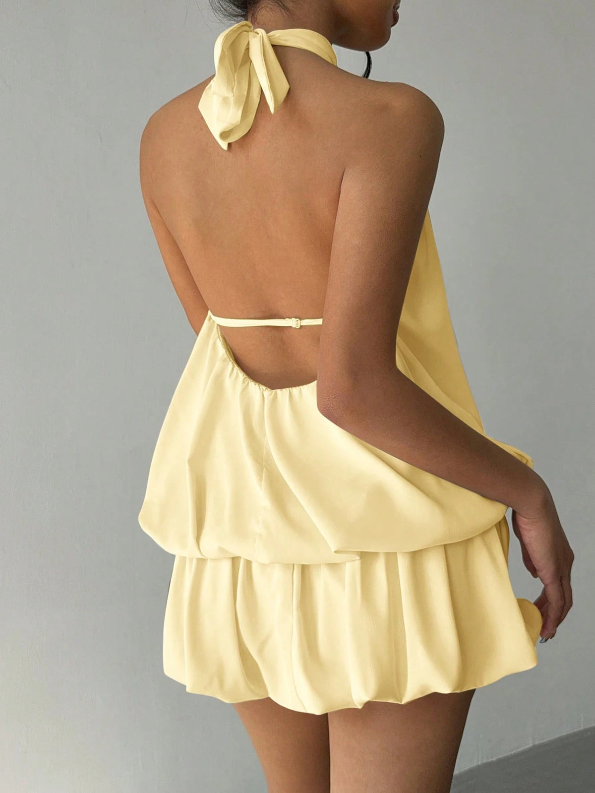 A model shown from the back wearing a pale yellow halter top with a tie-neck bow and a layered ruffled mini skirt, set against a plain gray background.