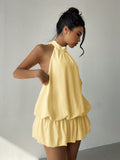 A woman wearing a pale yellow halter-neck romper with a ruffled tiered skirt, standing against a plain light gray wall.