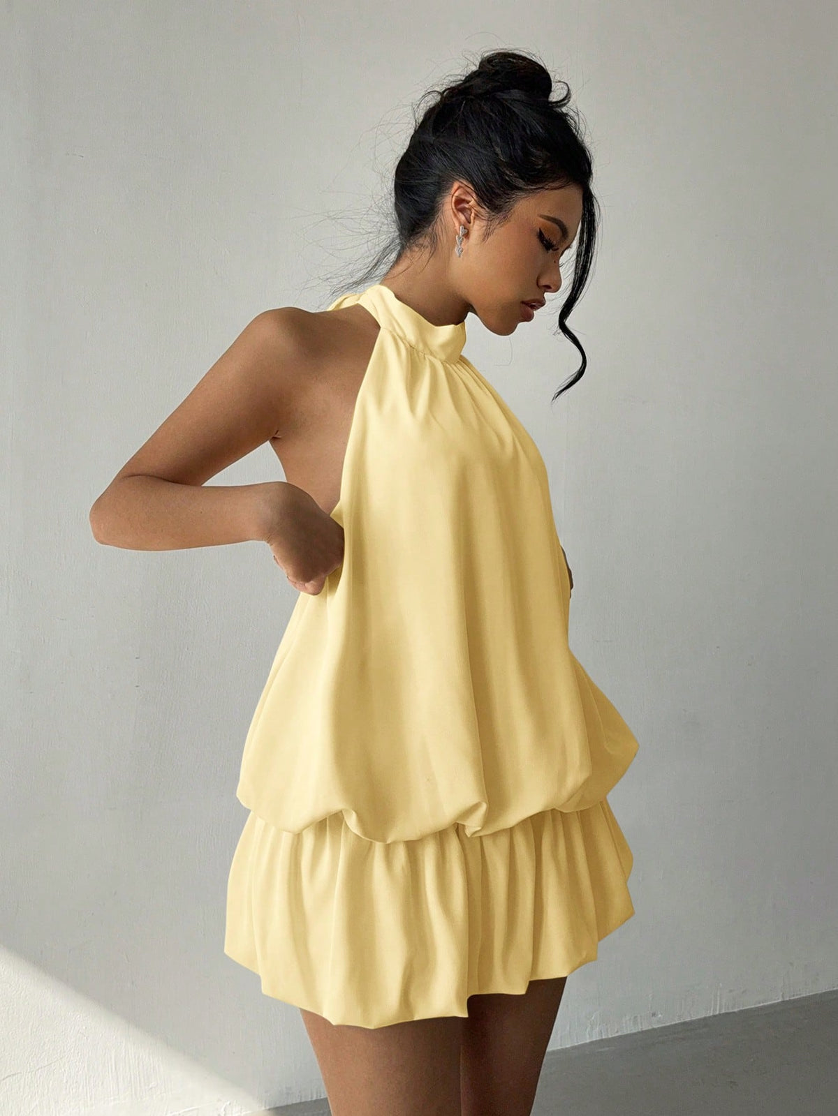 A woman wearing a pale yellow halter-neck romper with a ruffled tiered skirt, standing against a plain light gray wall.
