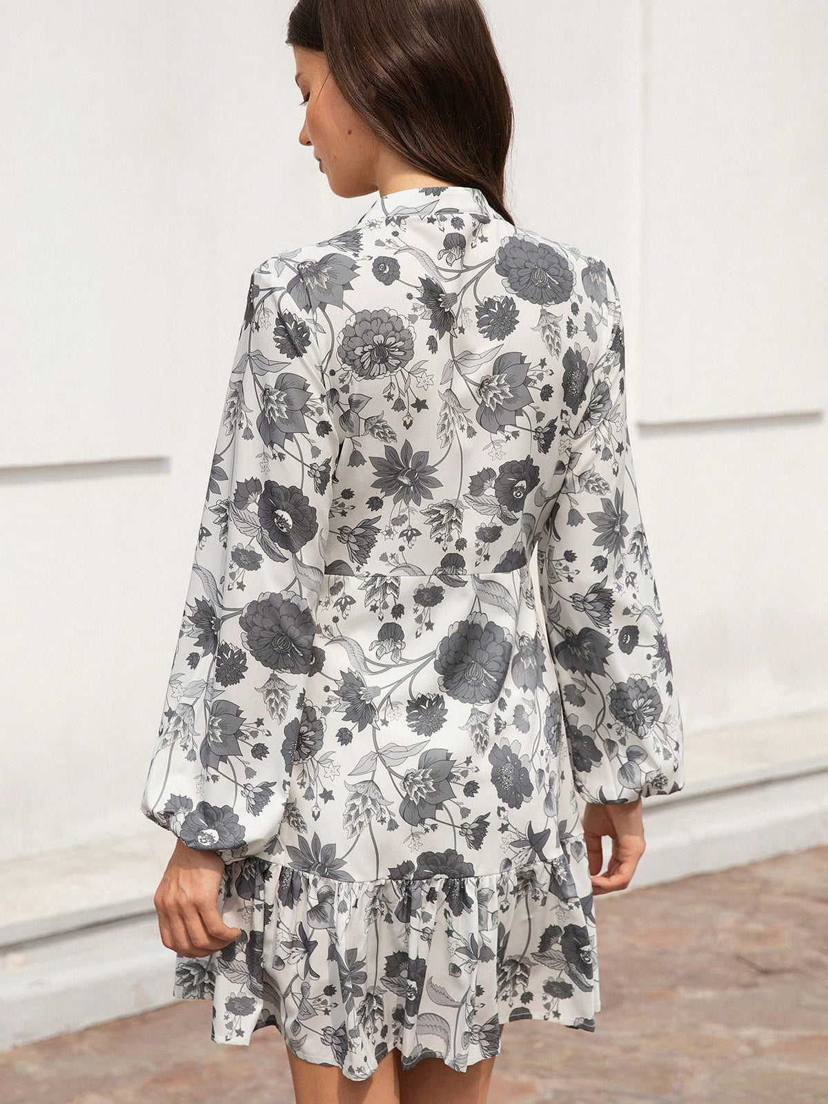 A woman shown from the back wearing a long-sleeve floral dress with a ruffled hem, featuring a black and gray floral pattern on a white background.