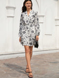 A woman models a long-sleeve white mini dress with a black floral print, standing outdoors against a white stucco wall with arches.