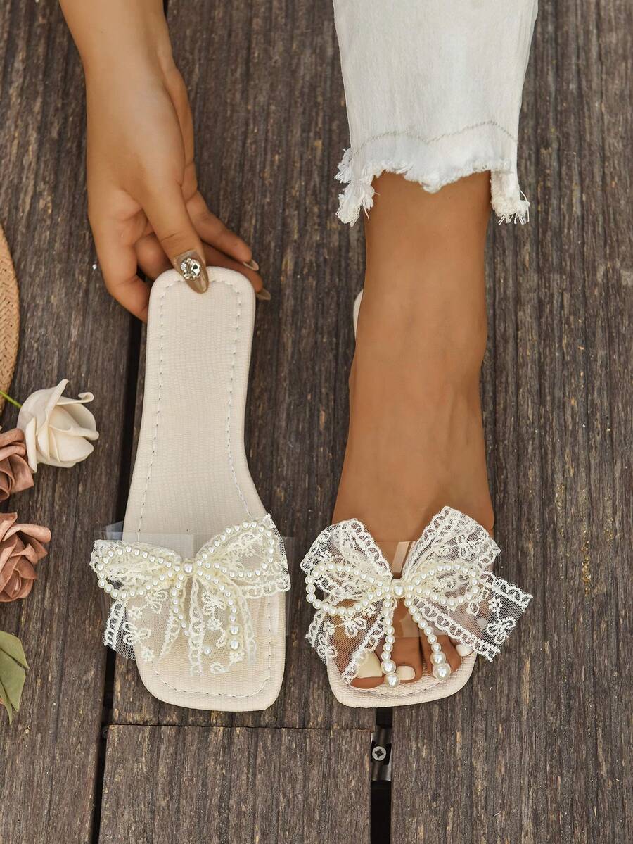 Cream lace bow flat slide sandal with pearl trim on wood deck