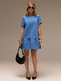 A woman modeling a short-sleeve light blue denim mini dress with a ruffled hem, styled with black pointed-toe heels, gold jewelry, sunglasses, and a black crescent handbag against a neutral studio backdrop.