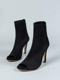 A pair of black knit ankle boots with open peep toes and slim high stiletto heels, photographed on a light gray studio background.