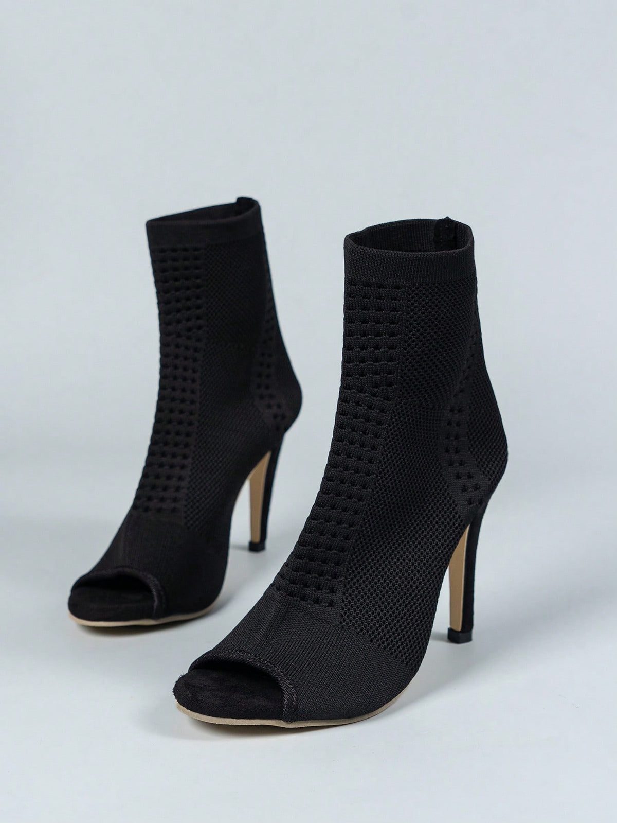 A pair of black knit ankle boots with open peep toes and slim high stiletto heels, photographed on a light gray studio background.