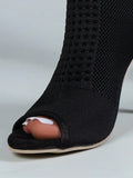 A close-up of a black open-toe peep-toe high heel shoe with a textured knit upper, shown on a foot with white toenail polish against a light gray background.