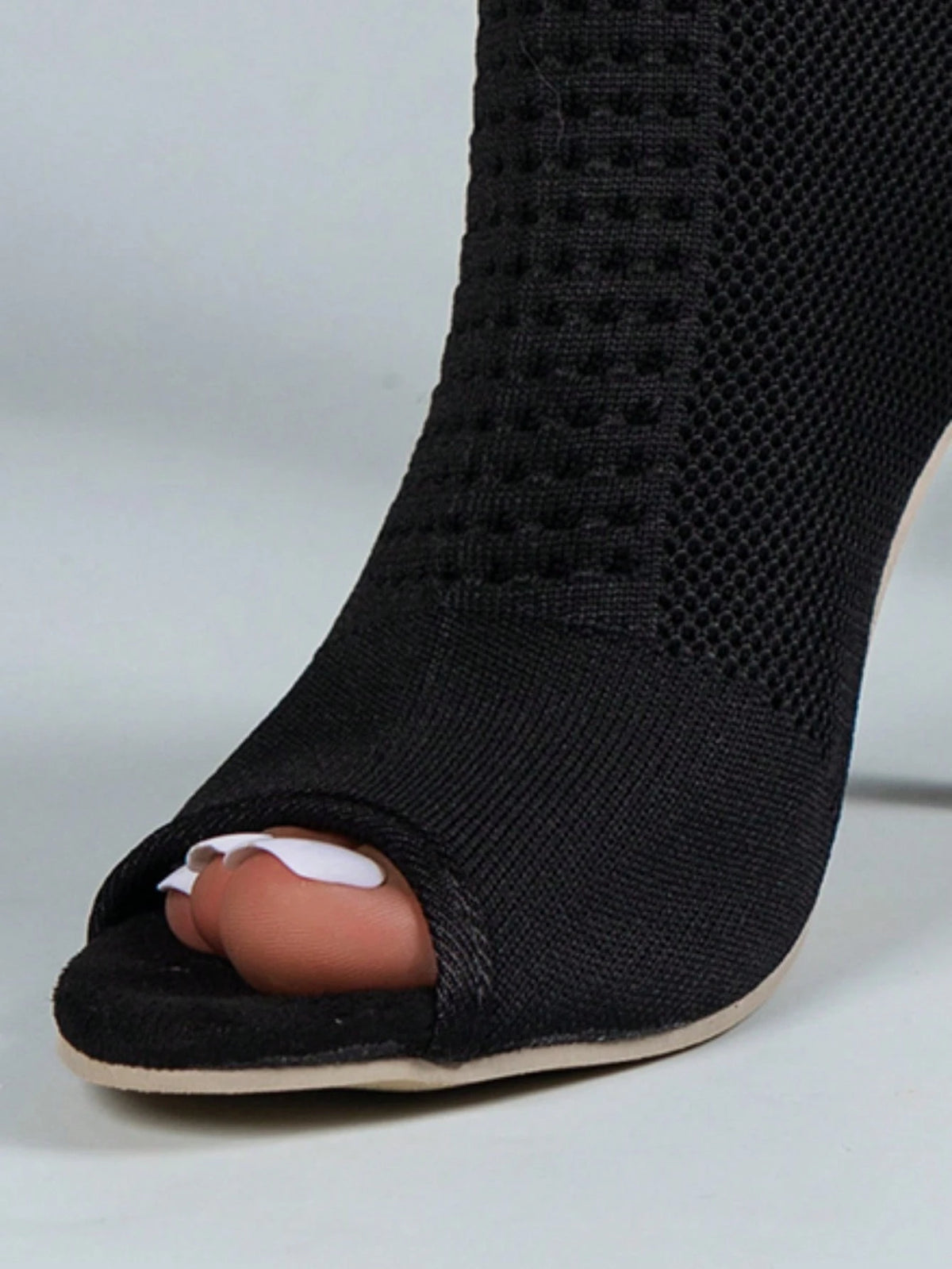 A close-up of a black open-toe peep-toe high heel shoe with a textured knit upper, shown on a foot with white toenail polish against a light gray background.