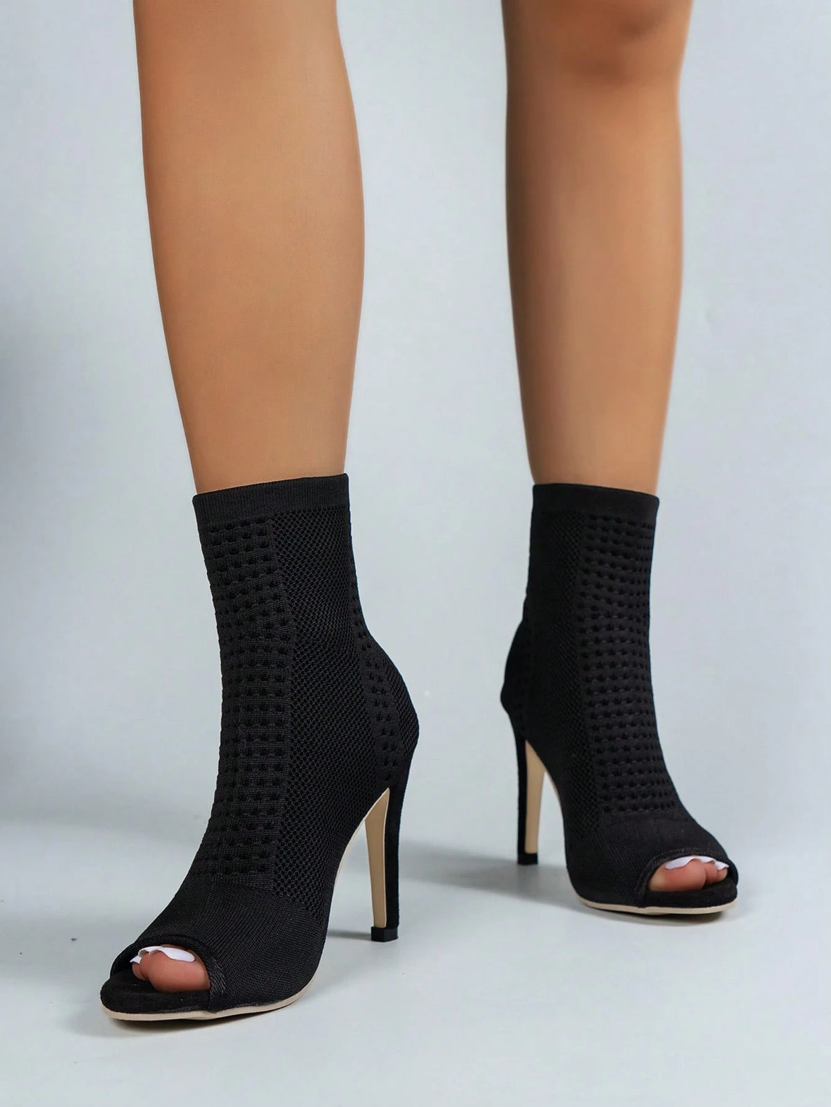 Black knit ankle boots with open-toe peep toes and high stiletto heels, shown on a model's feet against a light gray studio background.