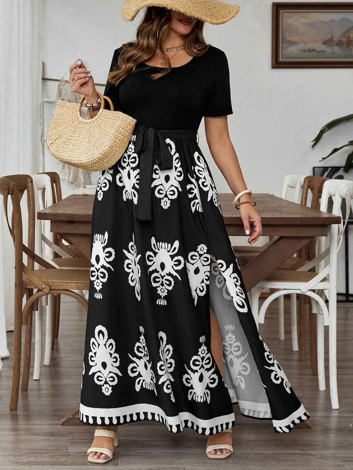 A woman wearing a black short-sleeve maxi dress with a white floral pattern, holding a woven straw bag and wearing a straw hat in a dining room setting.