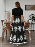 A woman viewed from behind wearing a black maxi dress with a solid black short-sleeve bodice and a flared skirt featuring large white floral and geometric patterns, standing in a dining room setting.