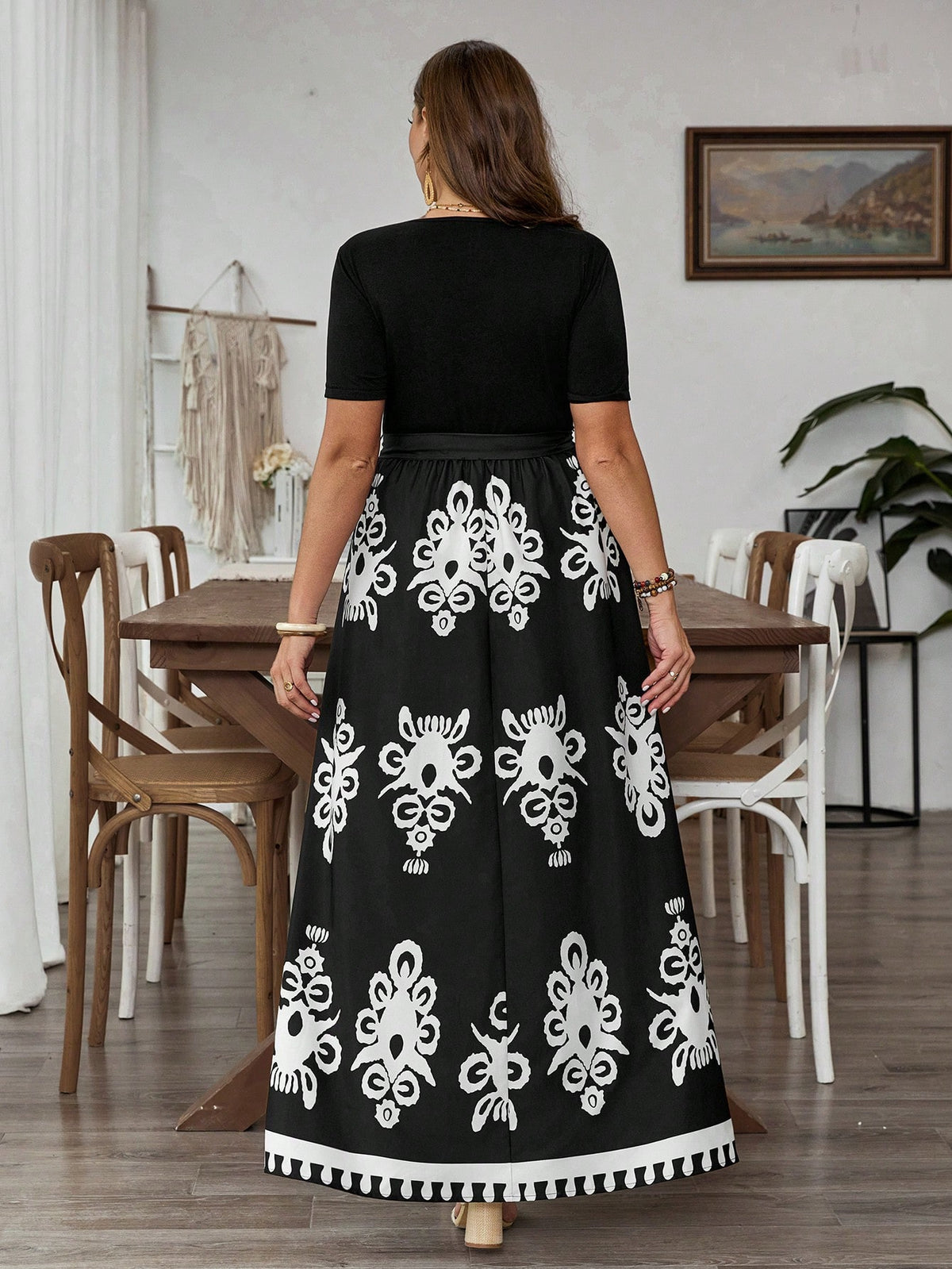 A woman viewed from behind wearing a black maxi dress with a solid black short-sleeve bodice and a flared skirt featuring large white floral and geometric patterns, standing in a dining room setting.