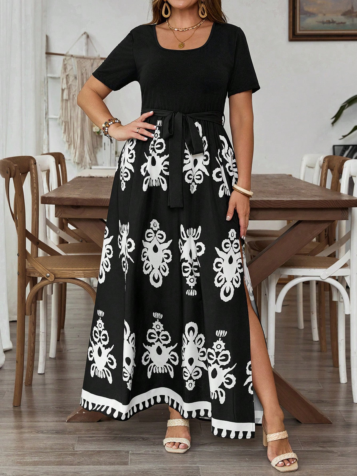 A woman models a black short-sleeve maxi dress with a white Mexican-style floral pattern and a side slit, standing in a dining room setting.
