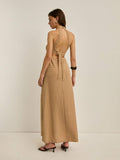 A woman wearing a beige halter-neck maxi dress with a textured geometric pattern, shown from the back against a plain off-white studio background.