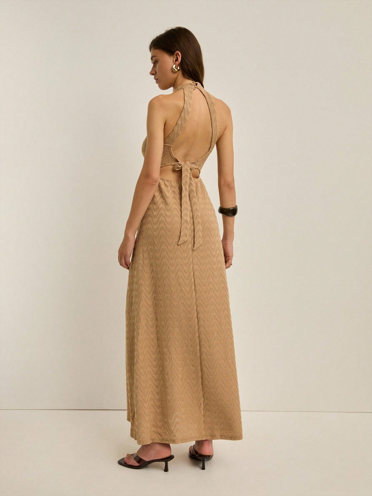 A woman wearing a beige halter-neck maxi dress with a textured geometric pattern, shown from the back against a plain off-white studio background.