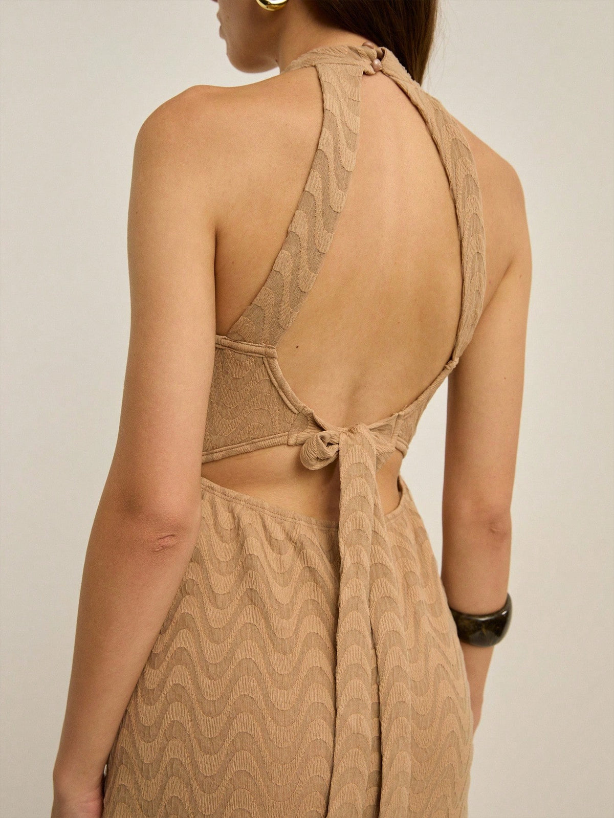 A woman shown from the back wearing a beige knit dress with a wavy pattern and an open back tied at the waist, photographed against a plain light background.