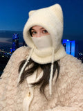 A woman wearing a cream-colored fuzzy cat-ear hooded face mask and a matching fluffy coat, photographed against a dark blue city skyline at night.
