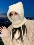 A person wearing a fluffy cream-colored knit hood with cat ears and a face covering, paired with a textured cream coat, photographed outdoors at dusk with a city skyline in the background.