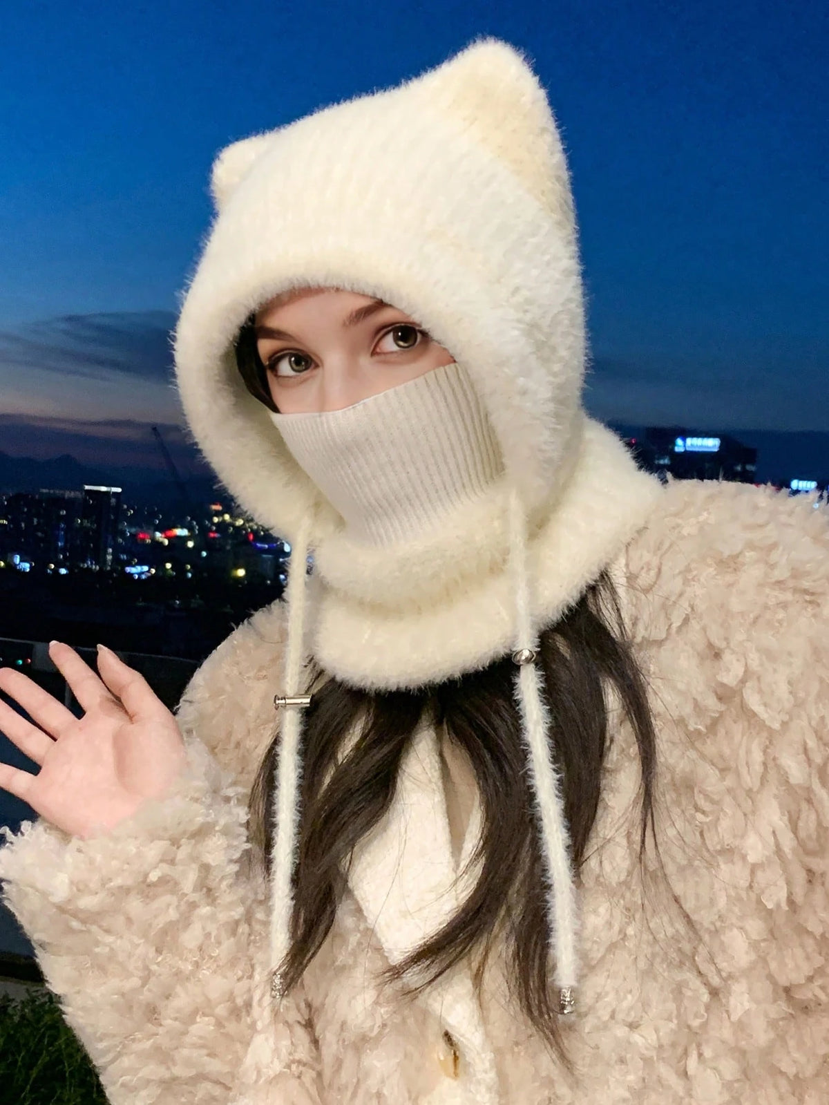 A person wearing a fluffy cream-colored knit hood with cat ears and a face covering, paired with a textured cream coat, photographed outdoors at dusk with a city skyline in the background.