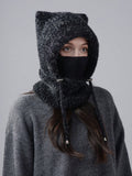 A person wearing a dark charcoal gray fuzzy knit hooded face mask with ear flaps and drawstrings, paired with a matching gray knit sweater, against a plain light gray studio background.