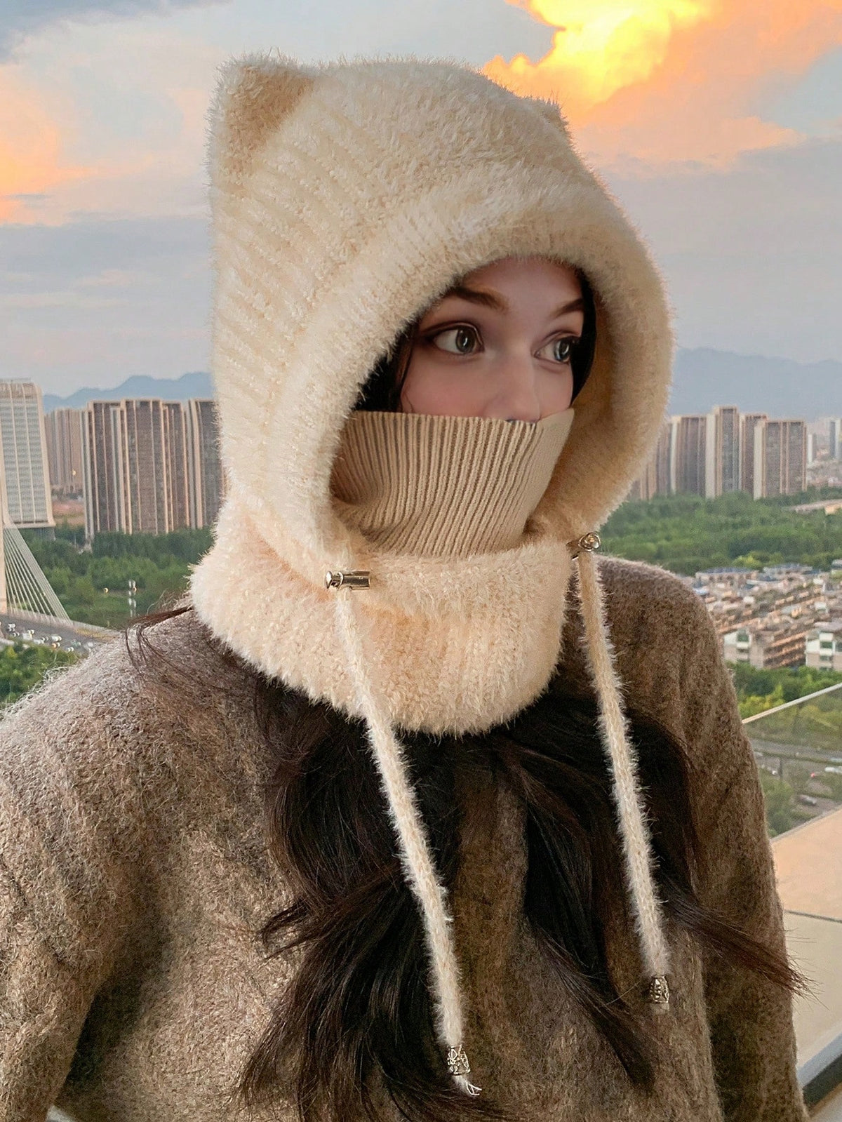 A woman wearing a cream-colored fuzzy knit hooded face mask with ear-like tips and a ribbed neck gaiter, photographed outdoors against a city skyline at sunset.