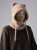 A person wearing a chunky knit winter hood with bear ears, a face mask, and a neck gaiter in a warm beige/tan color, layered over a dark brown sweater against a plain gray studio background.
