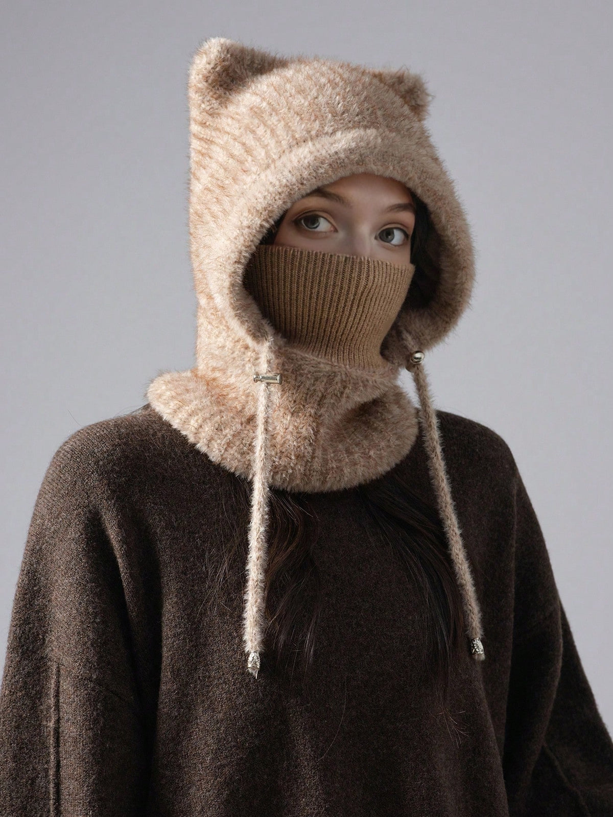 A person wearing a chunky knit winter hood with bear ears, a face mask, and a neck gaiter in a warm beige/tan color, layered over a dark brown sweater against a plain gray studio background.