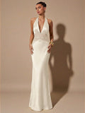A woman models a floor-length white halter gown with a deep V-neckline and a lace-embellished waistband, standing against a warm beige studio backdrop with a prominent shadow.