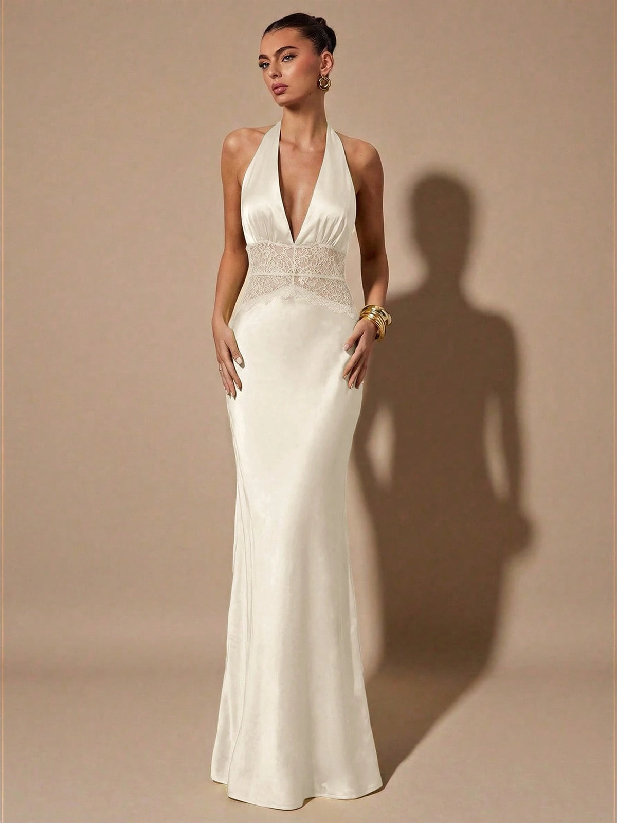 A woman models a floor-length white halter gown with a deep V-neckline and a lace-embellished waistband, standing against a warm beige studio backdrop with a prominent shadow.