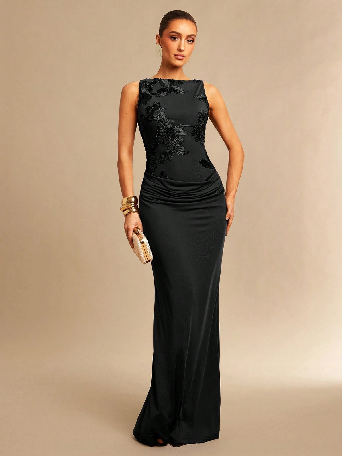 A woman modeling a sleeveless black floor-length gown with a fitted bodice, floral lace applique across the chest, and draped ruching at the waist, standing against a plain beige studio background.